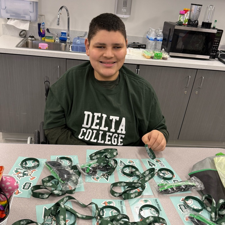 adult mae transition student sitting in a wheelchair at his desk smiles at the camera. piles of green delta college swag sit on the table in front of him  