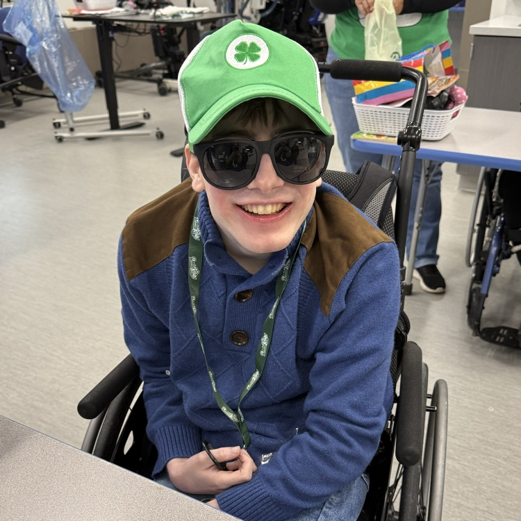 adult male transition student sitting in a wheelchair smiles at the camera he is wearing a green hat and green sunglasses for st Patrick’s day  