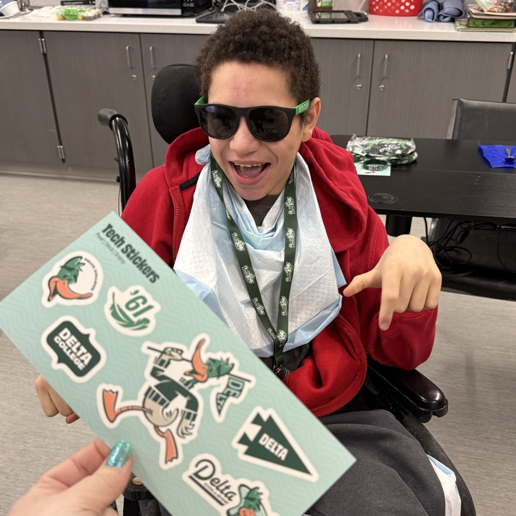 adult male transition student sits in a wheelchair smiling wearing green lanyard and sunglasses and holding a page of delta college stickers  