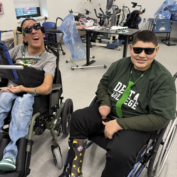 two adult male transition students sit in their wheelchairs smacking at the camera wearing green sunglasses and lanyards and ties to celebrate st Patrick’s day  