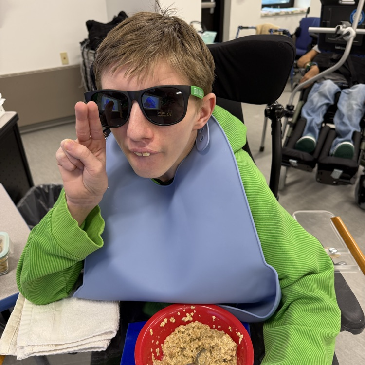 adult male transition student in a wheelchair gives the peace sign while wearing green shirt, sunglasses for st Patrick’s day  