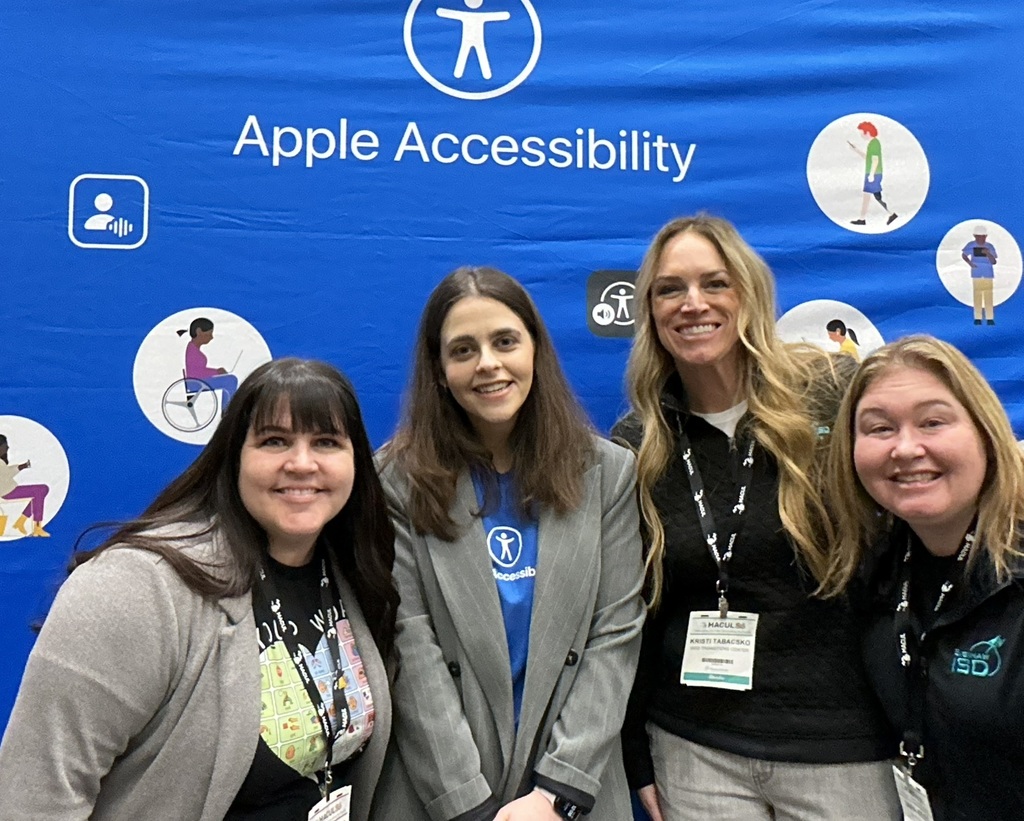 Four women smile together in front of a bright blue Apple Accessibility display at a conference. The backdrop features the words “Apple Accessibility” and several accessibility-themed icons and illustrations. All four are wearing conference lanyards, and the group appears to be posing for a celebratory photo at the event.