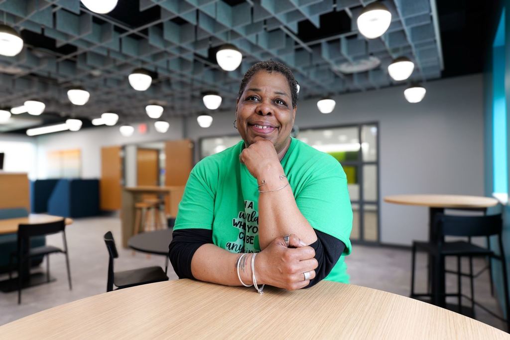 Person in green shirt sitting at a table with hand on chin, in a room with chairs and tables.