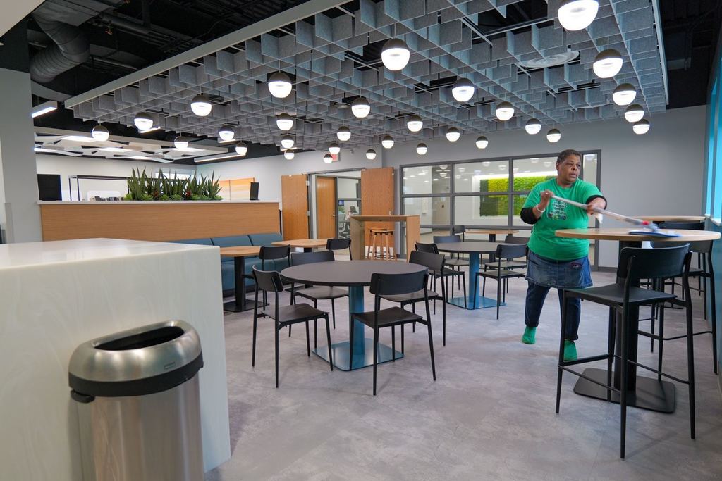 A person cleans a table in a dining area with round tables, chairs, and a trash can.