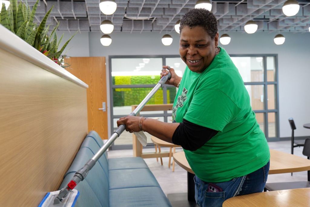A person in a green shirt and blue jeans uses a mop to clean a blue bench in a room.