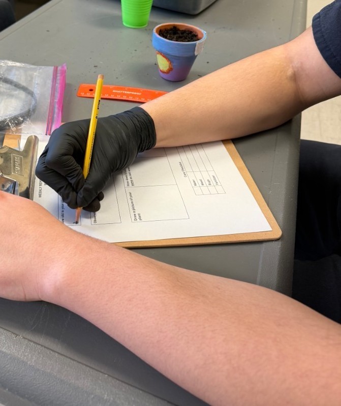 student writing with a gloved hand in pencil on a worksheet held by a clipboard next to an orange ruler and small, colorful planter full of soil