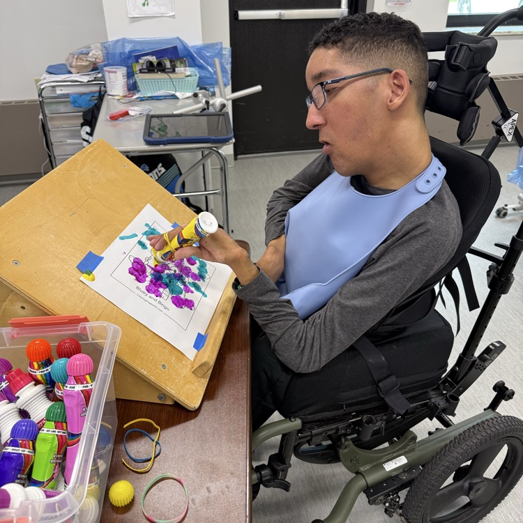 Young adult male wheelchair student uses physical accommodations of a slant board and grip holder to paint on a Mother’s Day coloring page in a classroom 