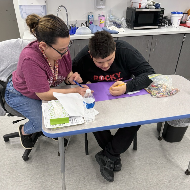 Young adult male wheelchair user student is assisted by a female para pro to write words on a Mother’s Day card