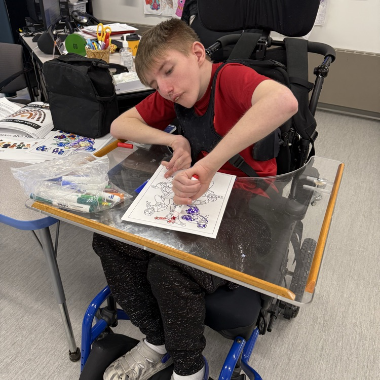 Young adult wheelchair user student uses a marker to color on a paper on his wheelchair tray in a classroom