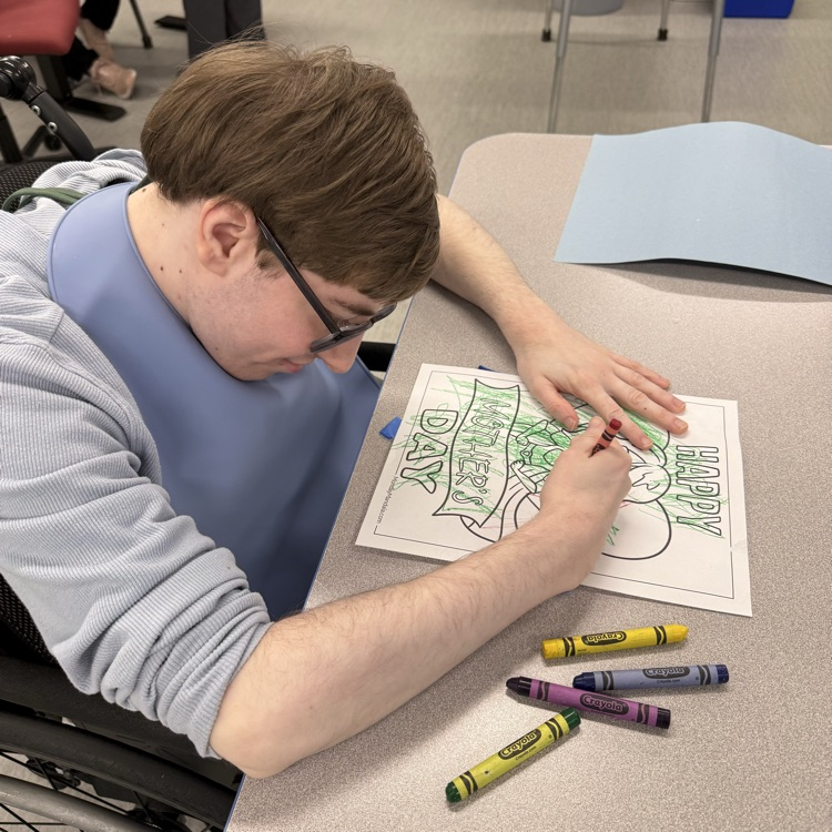 Young adult male wheelchair user student uses crayons to color on a coloring page on the table in front of him in a classroom