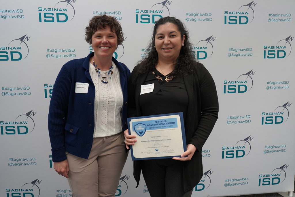 Two women, one holding a certificate, stand next to each other in front of a blue backdrop with white text.