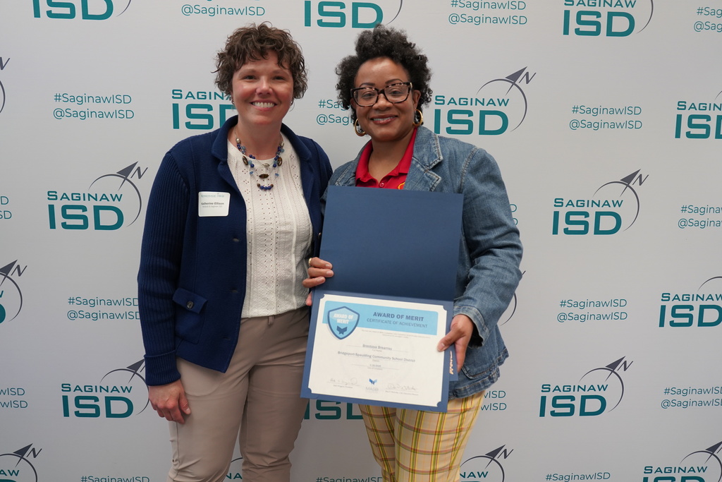 Two women stand by a backdrop with the text "ISD" and "Saginaw ISD" holding a framed certificate.