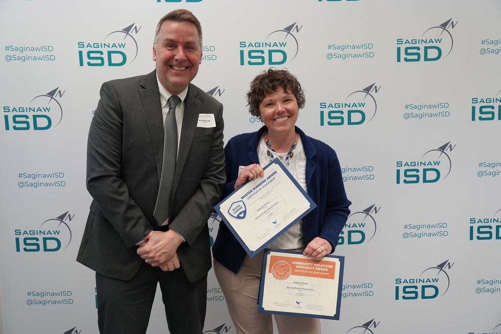 Two people hold certificates. A man in a suit, a woman in a blue blazer. They stand against a wall with a blue and white pattern.