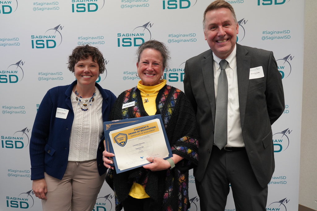 Three people stand by a banner with logos. The middle person holds an award certificate. The left person wears a jacket. The right wears a suit and tie.