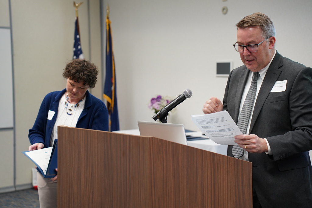 A podium with a microphone, papers, and a laptop. A man in a suit speaks while a woman stands behind him.