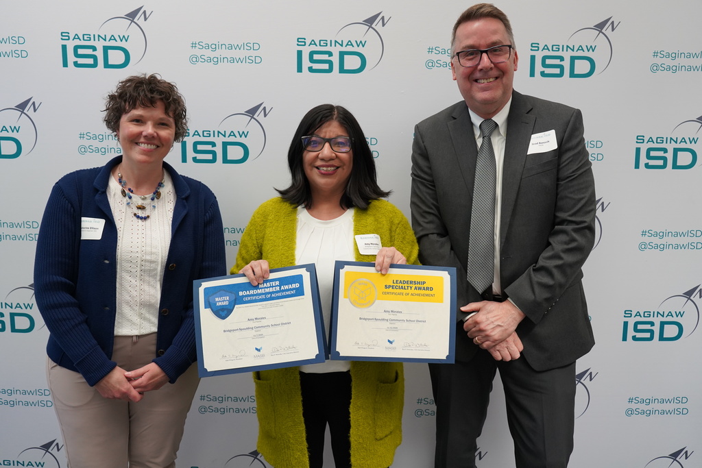 Three people stand together, smiling and holding certificates. A woman in blue and a woman in yellow hold certificates. A man in a suit stands between them.