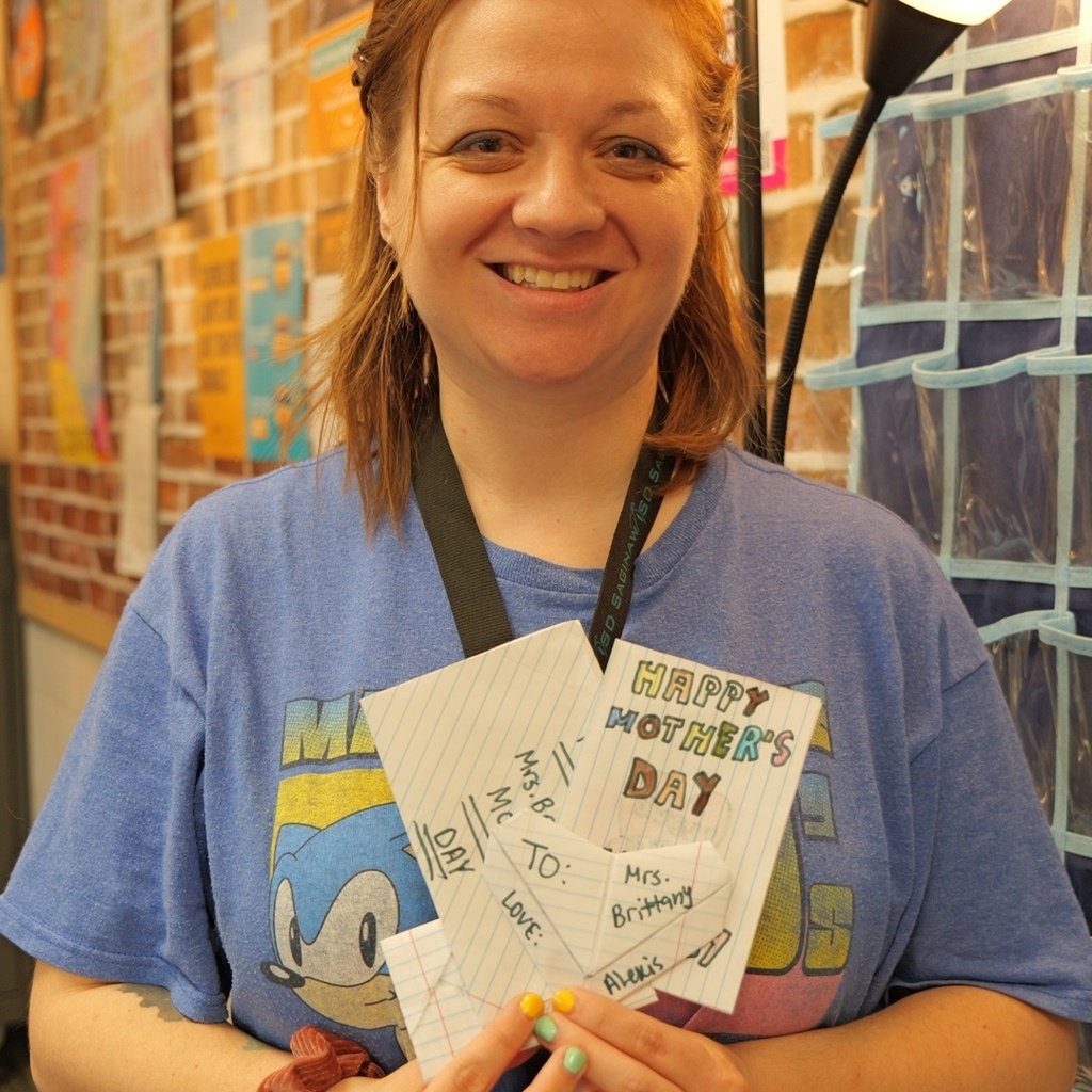 Person in a blue shirt with a graphic holds a card, smiling, with a brick wall in the background.