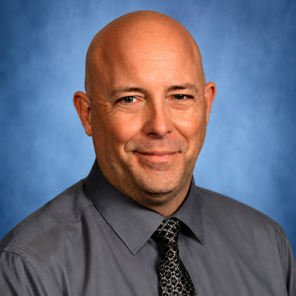 A man with a bald head, wearing a gray shirt and a patterned tie, smiling against a blue background.