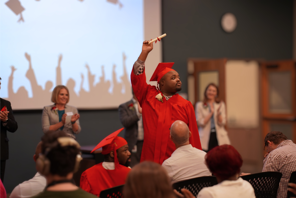 A graduate stands in red cap and gown, holding a diploma aloft, while others clap. Background features a projection of celebrating hands.