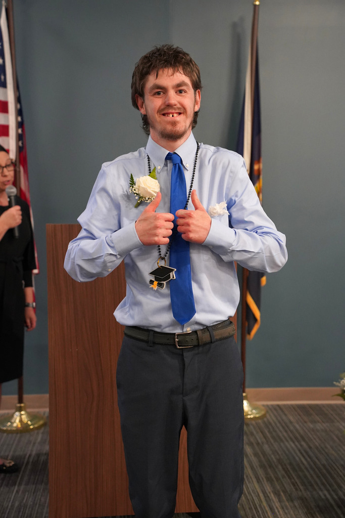 A person in a blue shirt and tie gives a thumbs up, standing in front of a podium and flags.