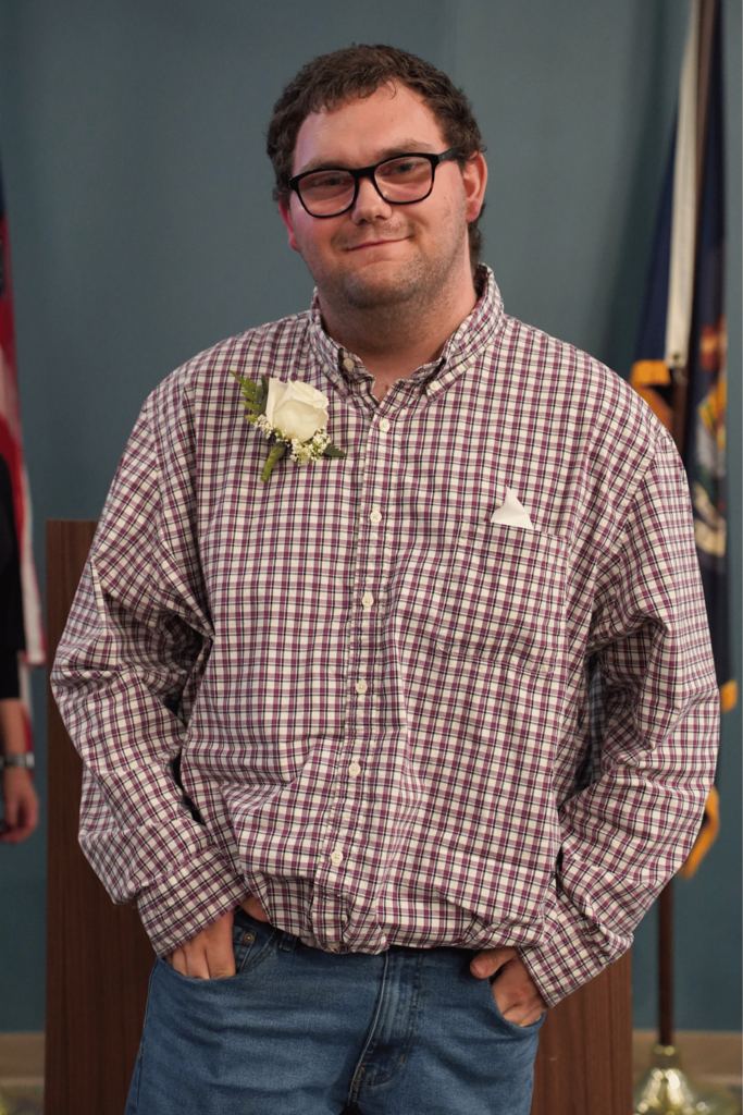 Man in plaid shirt and glasses with white flower on lapel, standing with hands in pockets. Behind him, a flag is visible.
