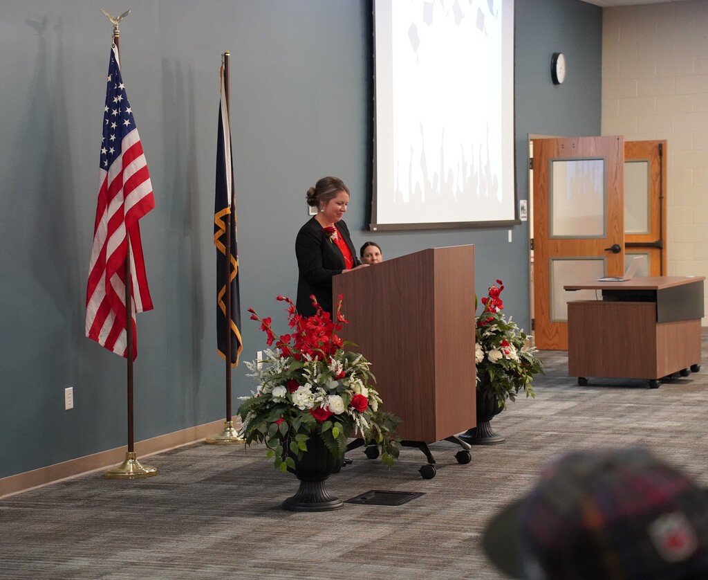 Person stands at podium in a room, next to an American flag and two flags on poles.