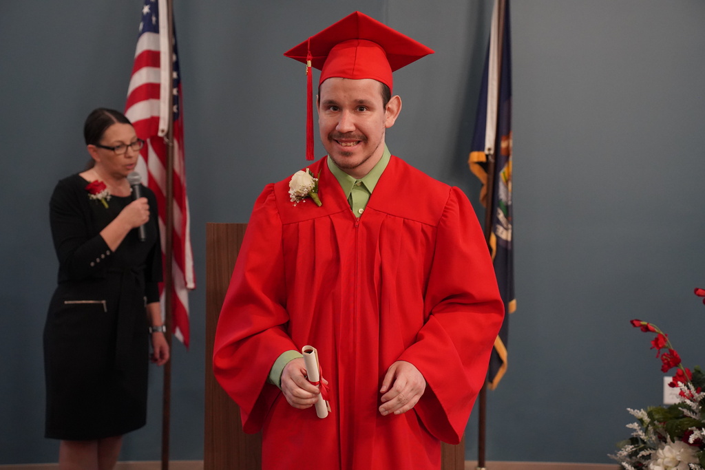 A man in a red graduation gown and cap stands in front of a podium, holding a diploma. Behind him, a woman with glasses holds a microphone.