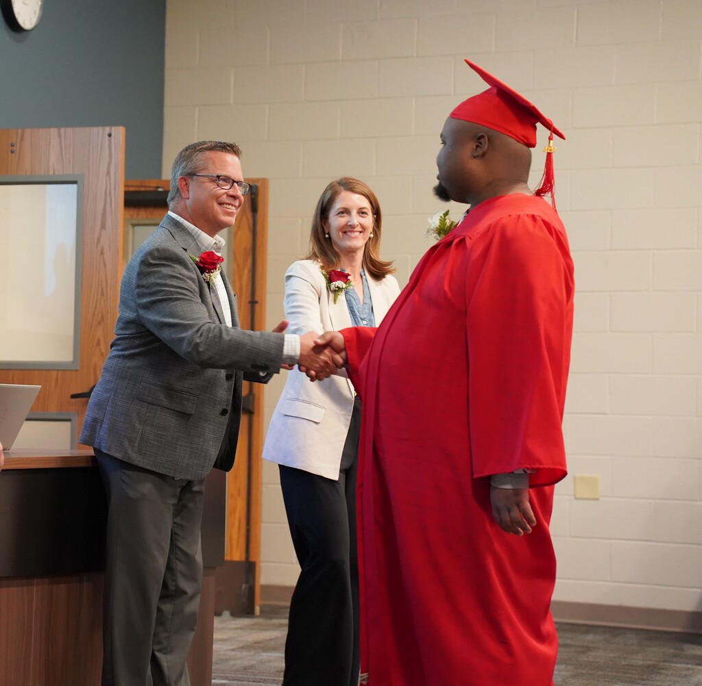 Man in cap and gown shakes hands with another man in a suit, near a woman in a blazer.
