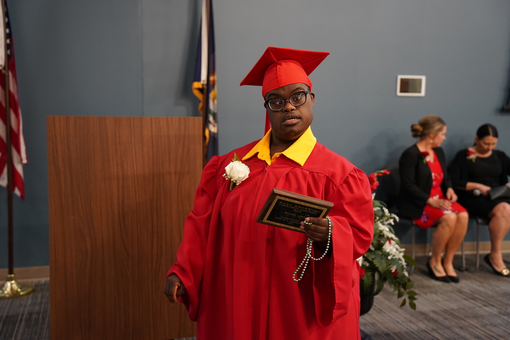Person in red cap and gown holds a plaque, standing before a podium. Behind them, two women sit and watch.