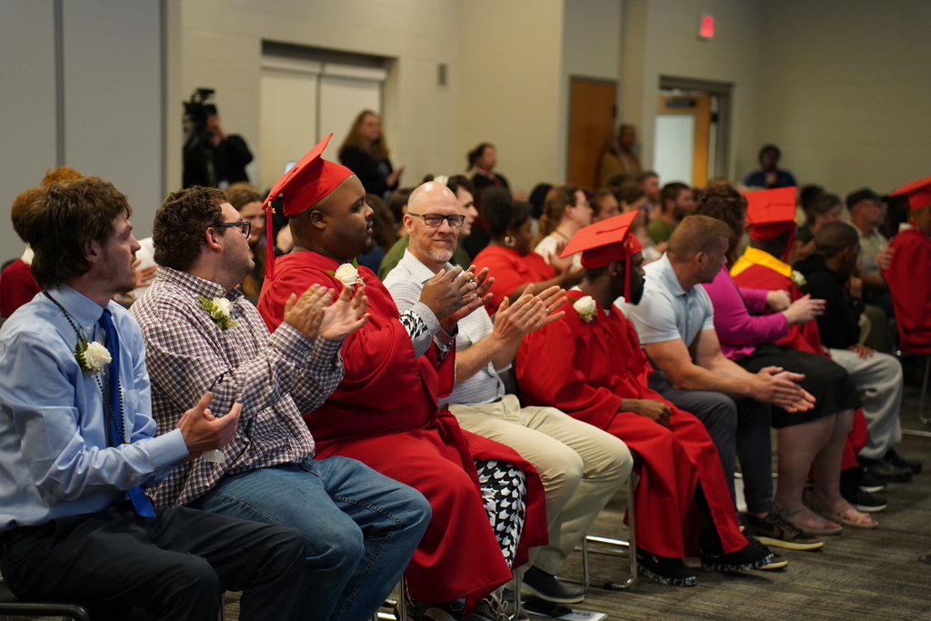 Group of people in red caps and gowns clapping in an auditorium. Some seated, others standing behind.
