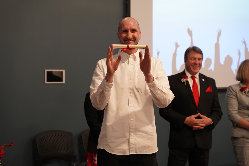 Man in white shirt holds a rolled paper, while standing before two other people in a room with a projector screen.