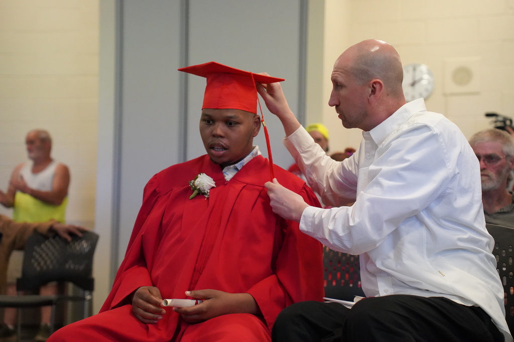 A man helps a person in a red graduation gown adjust their cap during a ceremony.