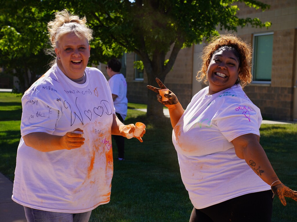 Two women in white t-shirts pose with hands covered in paint. One shows a peace sign. Background includes a grassy area and a building with windows.