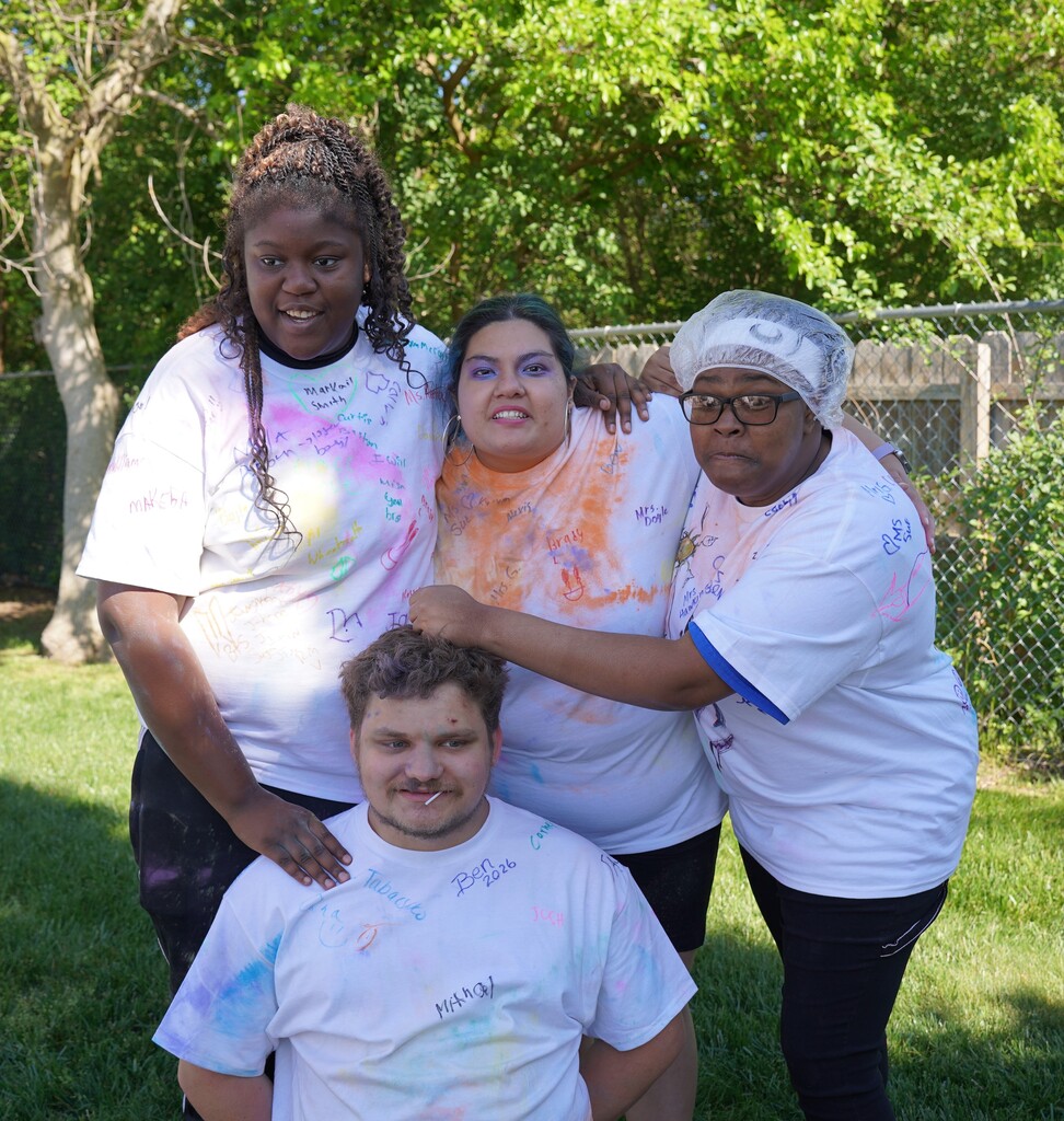 Four individuals, two women and two men, stand in a park with trees. They wear white shirts stained with colors. One person sits in front.