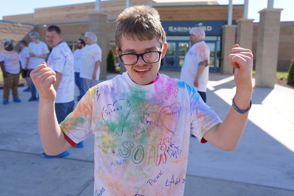 Boy wearing tie-dye shirt with "SOAR" and many signatures, smiles, and raises both arms. People in white shirts stand behind.