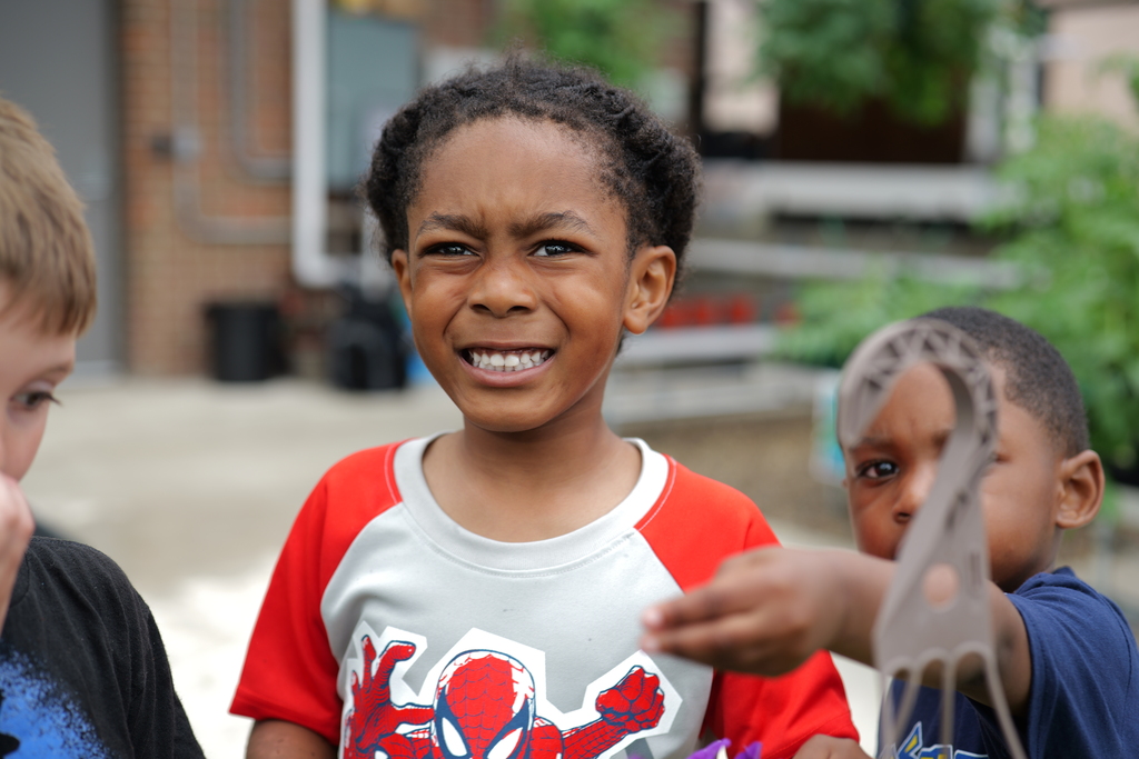 Three children in front of a building. One boy wears a red-white shirt with a spider-like design, another boy looks on.