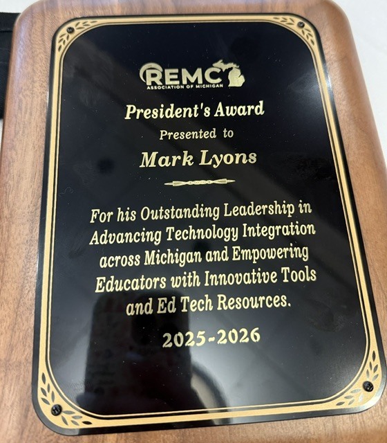 A wooden plaque with a black and gold engraved plate from the REMC Association of Michigan. The plaque reads: "President's Award Presented to Mark Lyons For his Outstanding Leadership in Advancing Technology Integration across Michigan and Empowering Educators with Innovative Tools and Ed Tech Resources. 2025–2026." The plaque recognizes Mark Lyons for his leadership in supporting educators and advancing educational technology throughout Michigan.