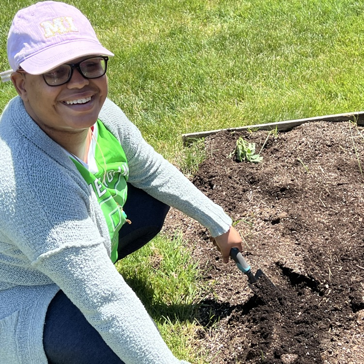 Person wearing a purple hat, glasses, and a gray sweater, kneeling and holding a tool over a garden bed.