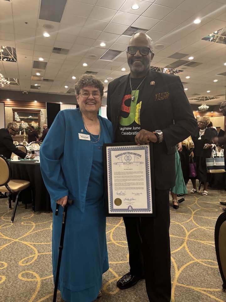 Man and woman holding certificate at event. The woman has a cane. Bright overhead lights.