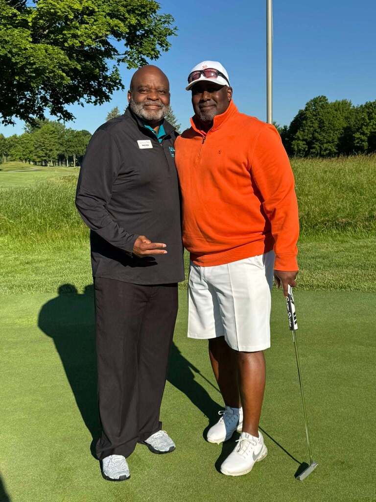 Two men standing on a golf course, one holding a club, smiling and posing for a photo.