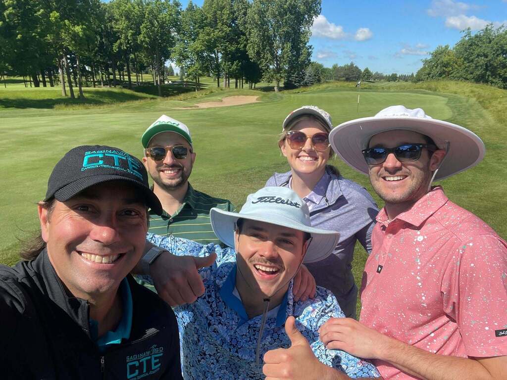 Five individuals wearing hats and sunglasses pose for a photo in a golf course with lush green grass.