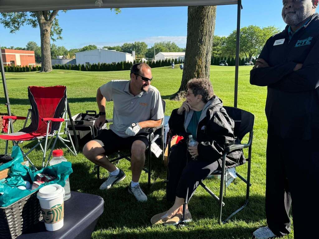 Three individuals sit and stand in a shaded area of a grassy field, with a table of refreshments nearby.