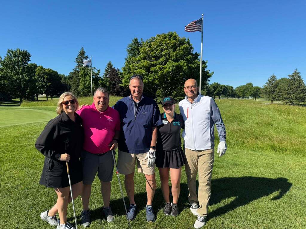 Five people stand on a golf course with a flagpole behind them. Two women and three men pose for a photo.