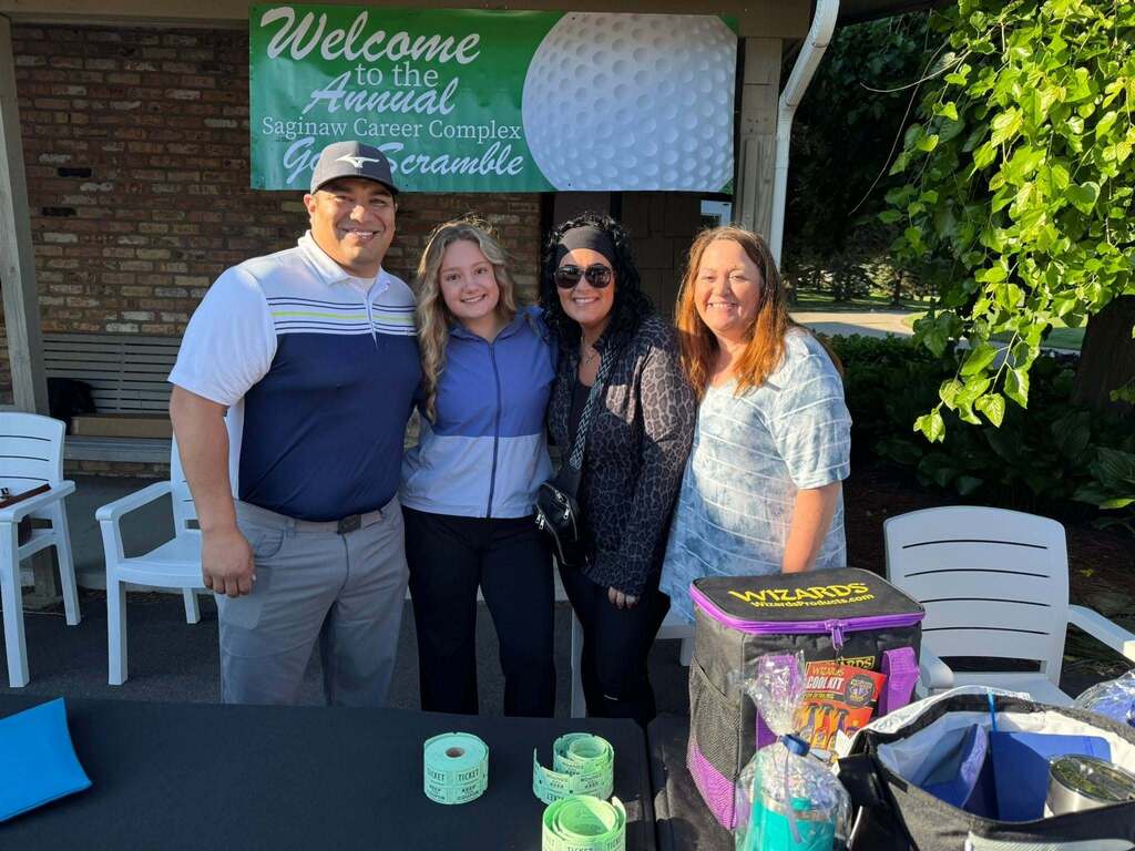 Four people stand in front of a golf event booth with a banner. Two women wear sunglasses and jackets.