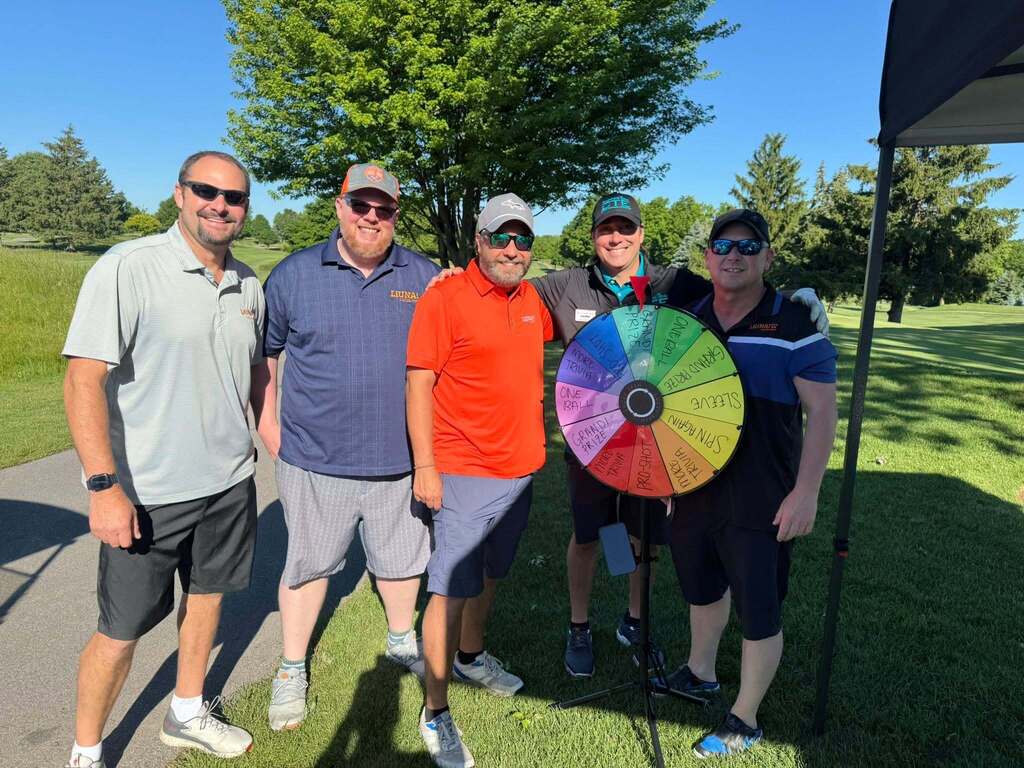 Five people pose for a picture on a golf course, with one holding a large, colorful wheel.