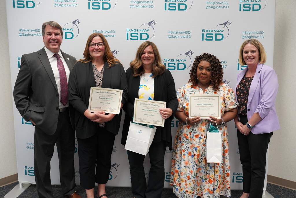 Five people, three women and a man, stand in front of a wall with a logo. They hold certificates and smile.