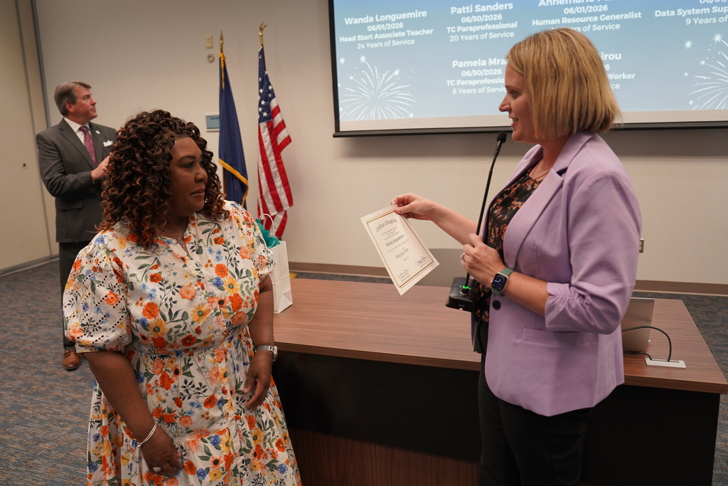 Two women in a conference room with a podium, flag, and screen behind them. One woman holds a paper.