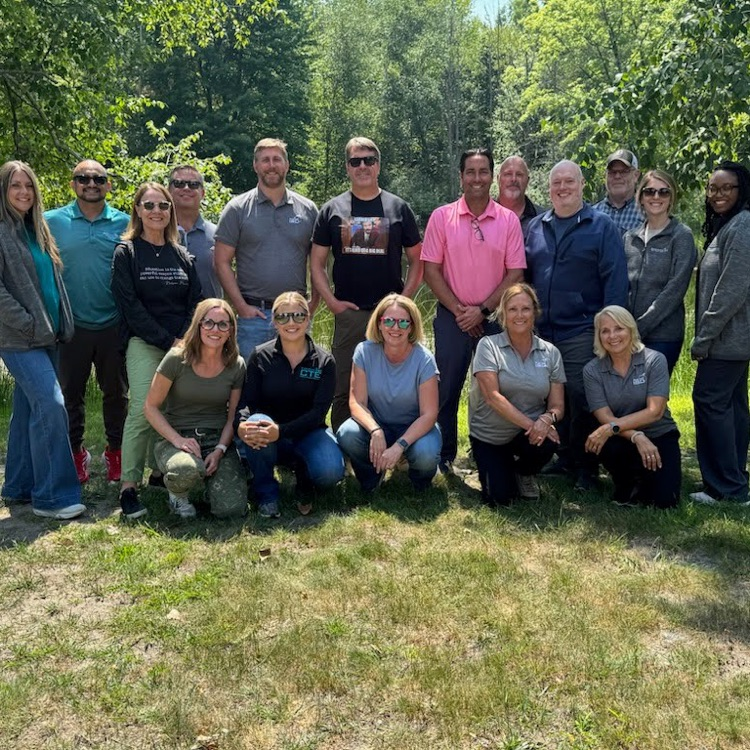 A group of people in casual clothing are standing and kneeling in a grassy area with trees.