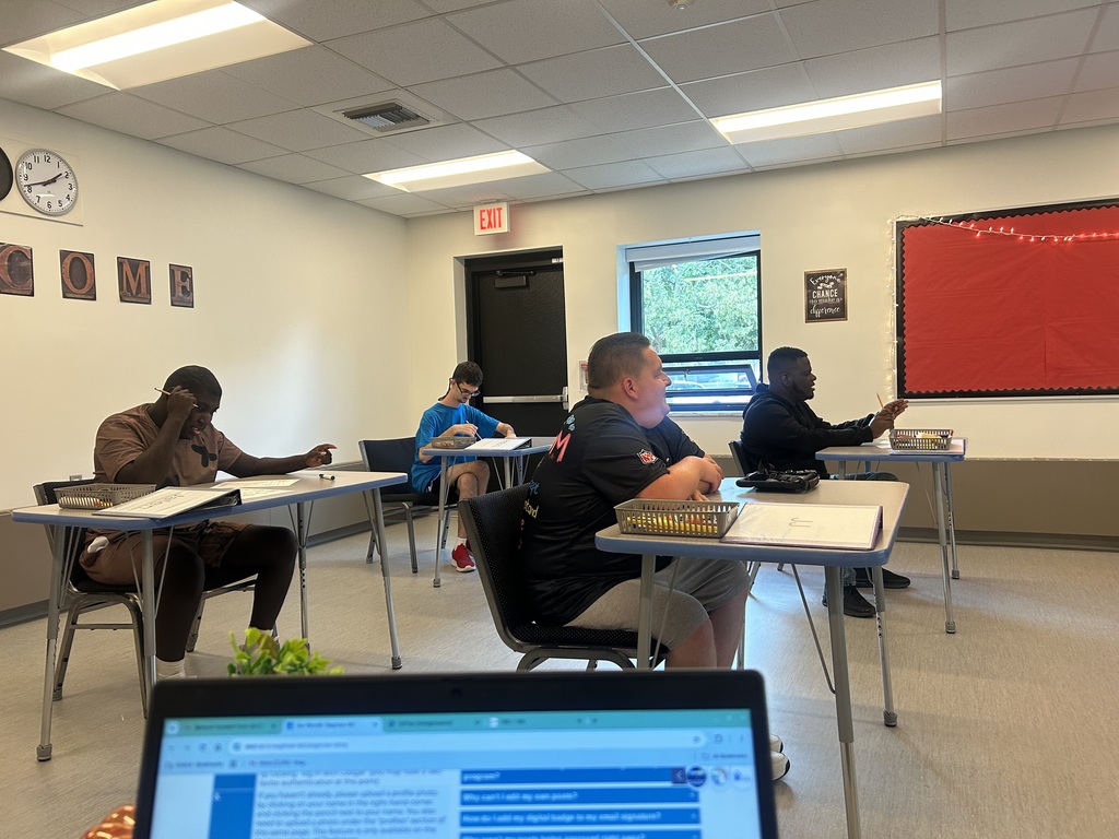 A classroom with desks, chairs, and five people working on papers. A clock, red board, and windows are visible.