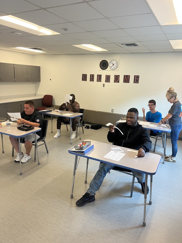 Several people seated at tables in a classroom. One person is seated in front, another is standing, and three people are seated in the background.